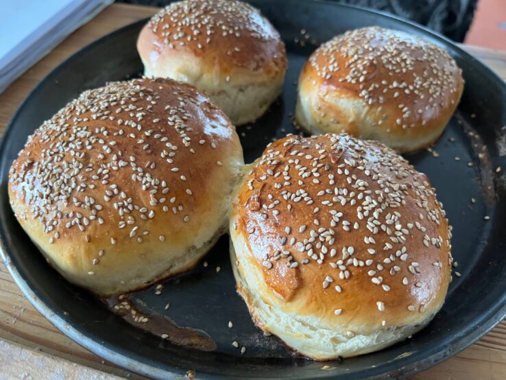Vier frisch gebackene Burger Buns mit goldbrauner Kruste und Sesam auf schwarzem Backblech