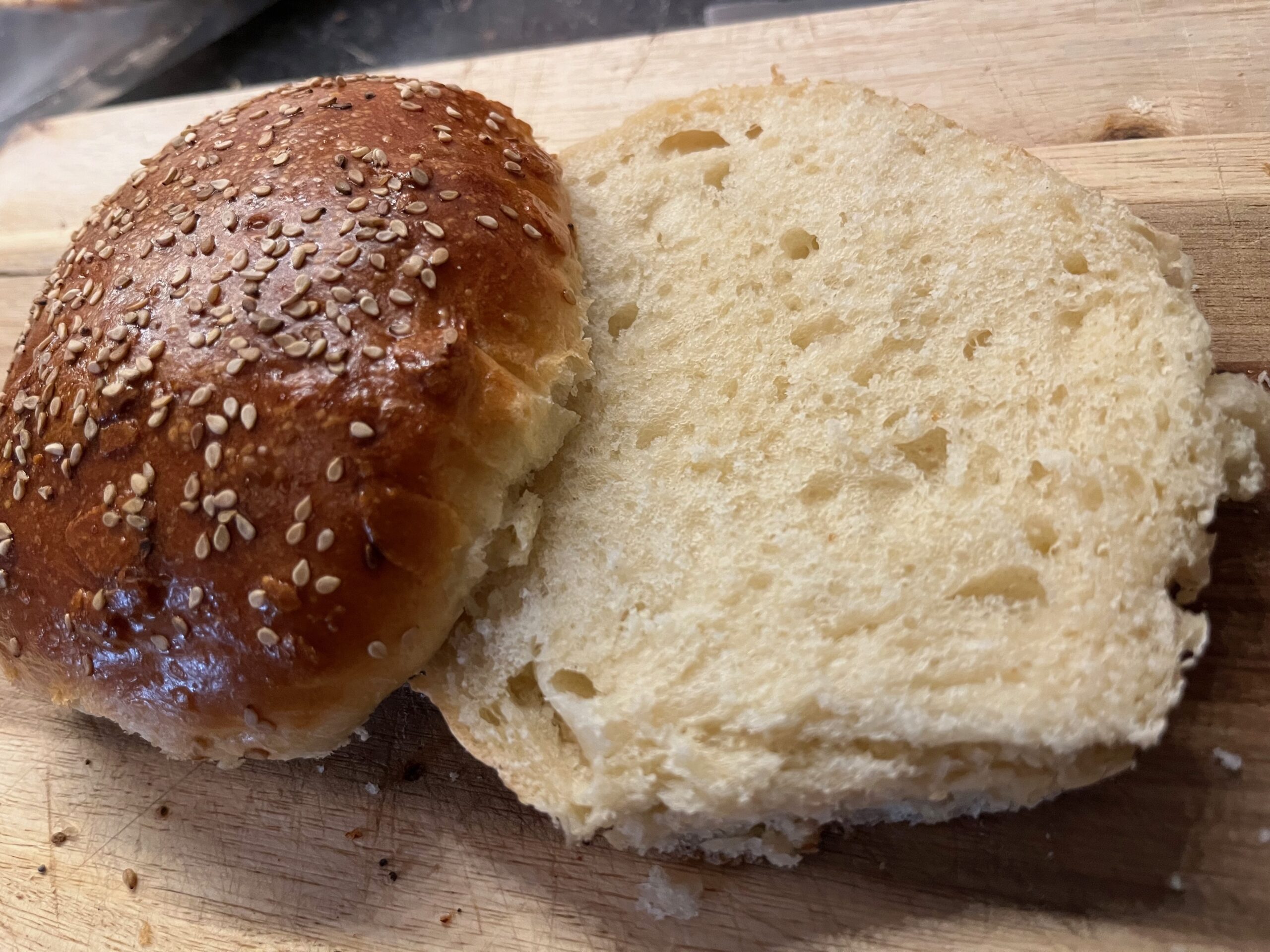 Aufgeschnittenes selbst gebackenes Burger Bun mit luftig-weicher Krume und glänzender Sesamkruste auf Holzbrett
