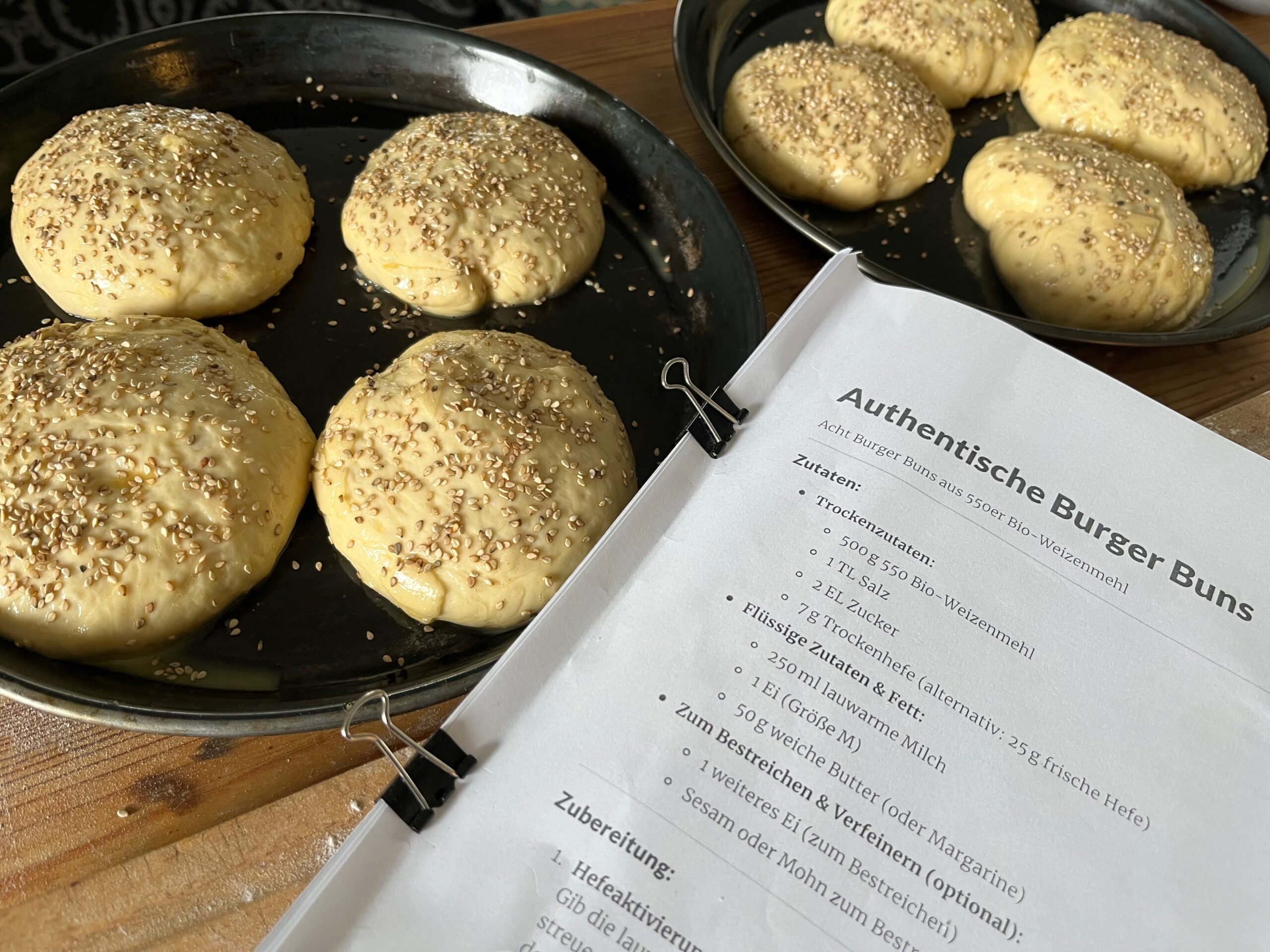 Rohe Burger-Bun-Teiglinge mit Sesam bestreut auf Backblech neben ausgedrucktem Rezept für authentische Burger Buns